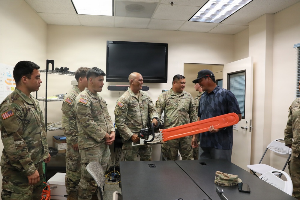 A group of U.S. Army soldiers in MultiCam/OCP camouflage uniforms are gathered in an indoor briefing or office room with a civilian instructor or demonstrator. The civilian is holding an orange-bladed chainsaw, apparently conducting a demonstration or training session. The atmosphere appears informal and engaged, with several soldiers smiling.