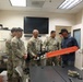 A group of U.S. Army soldiers in MultiCam/OCP camouflage uniforms are gathered in an indoor briefing or office room with a civilian instructor or demonstrator. The civilian is holding an orange-bladed chainsaw, apparently conducting a demonstration or training session. The atmosphere appears informal and engaged, with several soldiers smiling.