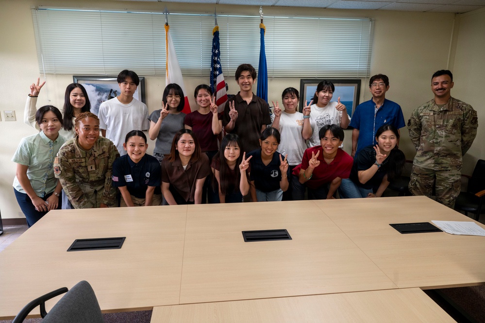 A group of approximately 20 people pose for a group photo in what appears to be a conference or briefing room. The group includes two individuals in U.S. military uniforms (camouflage/ACU pattern) alongside civilians, many of whom are making peace/victory hand signs. Three flags are visible in the background, including the U.S. flag, a Japanese flag, and what appears to be a U.S. Air Force flag.