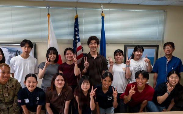 A group of approximately 20 people pose for a group photo in what appears to be a conference or briefing room. The group includes two individuals in U.S. military uniforms (camouflage/ACU pattern) alongside civilians, many of whom are making peace/victory hand signs. Three flags are visible in the background, including the U.S. flag, a Japanese flag, and what appears to be a U.S. Air Force flag.
