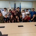 A group of approximately 20 people pose for a group photo in what appears to be a conference or briefing room. The group includes two individuals in U.S. military uniforms (camouflage/ACU pattern) alongside civilians, many of whom are making peace/victory hand signs. Three flags are visible in the background, including the U.S. flag, a Japanese flag, and what appears to be a U.S. Air Force flag.
