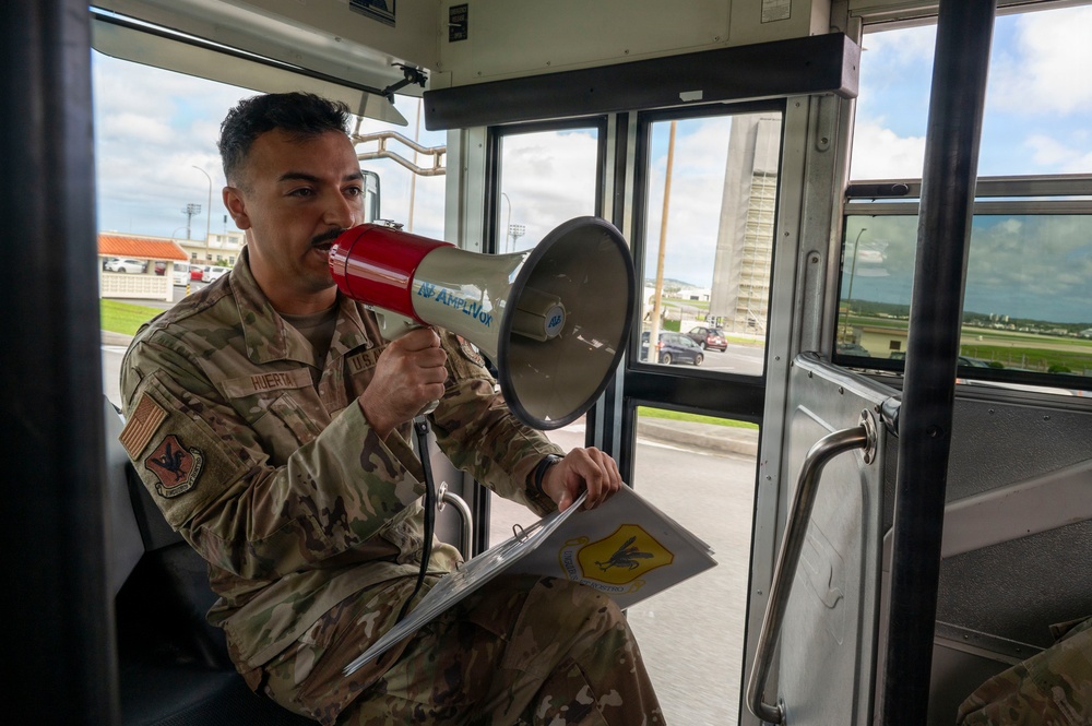 A U.S. Air Force service member named Huerta is seated inside a military bus, speaking into a red and gray megaphone branded 'Amplivox'. He is holding a clipboard with what appears to be a unit patch or insignia visible on the documents. The setting appears to be a military installation, likely an air base, visible through the bus windows.