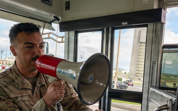 A U.S. Air Force service member named Huerta is seated inside a military bus, speaking into a red and gray megaphone branded 'Amplivox'. He is holding a clipboard with what appears to be a unit patch or insignia visible on the documents. The setting appears to be a military installation, likely an air base, visible through the bus windows.