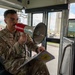 A U.S. Air Force service member named Huerta is seated inside a military bus, speaking into a red and gray megaphone branded 'Amplivox'. He is holding a clipboard with what appears to be a unit patch or insignia visible on the documents. The setting appears to be a military installation, likely an air base, visible through the bus windows.