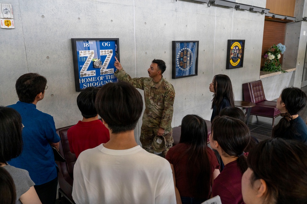 A U.S. Air Force service member in OCP (Operational Camouflage Pattern) uniform is briefing a group of civilians, likely Asian visitors or students, in what appears to be a building lobby or hallway. The soldier is pointing to a framed display on the wall that reads '18th OG - Home of the Shoguns,' which is associated with the 18th Operations Group, likely at Kadena Air Base in Japan. Several framed military unit insignia and shadow boxes are mounted on the concrete wall behind him.