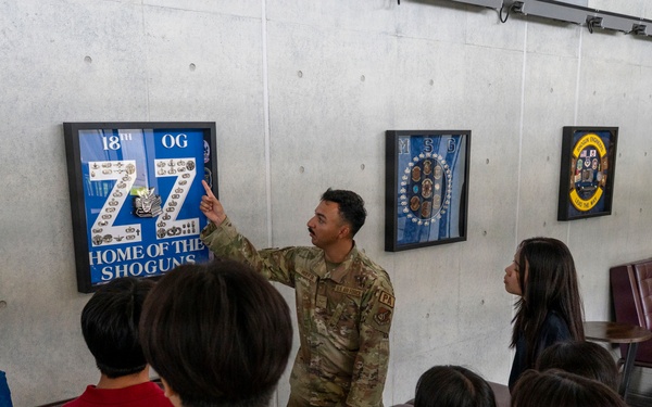 A U.S. Air Force service member in OCP (Operational Camouflage Pattern) uniform is briefing a group of civilians, likely Asian visitors or students, in what appears to be a building lobby or hallway. The soldier is pointing to a framed display on the wall that reads '18th OG - Home of the Shoguns,' which is associated with the 18th Operations Group, likely at Kadena Air Base in Japan. Several framed military unit insignia and shadow boxes are mounted on the concrete wall behind him.