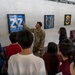 A U.S. Air Force service member in OCP (Operational Camouflage Pattern) uniform is briefing a group of civilians, likely Asian visitors or students, in what appears to be a building lobby or hallway. The soldier is pointing to a framed display on the wall that reads '18th OG - Home of the Shoguns,' which is associated with the 18th Operations Group, likely at Kadena Air Base in Japan. Several framed military unit insignia and shadow boxes are mounted on the concrete wall behind him.