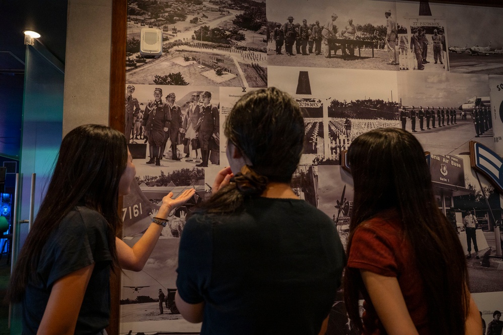 Three young people, viewed from behind, are examining a large wall collage of black-and-white historical military photographs in what appears to be a museum or exhibit space. The photos depict various military scenes including formations, ceremonies, and what appears to be an air force base. One person is pointing at a specific photograph on the wall.