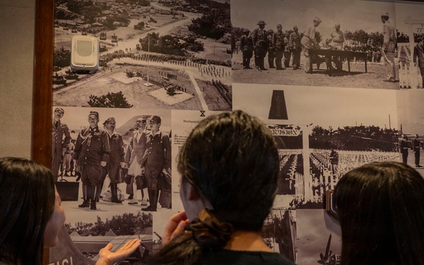 Three young people, viewed from behind, are examining a large wall collage of black-and-white historical military photographs in what appears to be a museum or exhibit space. The photos depict various military scenes including formations, ceremonies, and what appears to be an air force base. One person is pointing at a specific photograph on the wall.