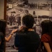 Three young people, viewed from behind, are examining a large wall collage of black-and-white historical military photographs in what appears to be a museum or exhibit space. The photos depict various military scenes including formations, ceremonies, and what appears to be an air force base. One person is pointing at a specific photograph on the wall.