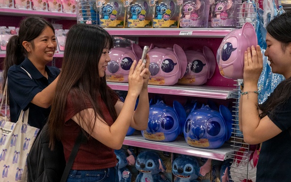 Three young women are shopping in a toy store, gathered around a display shelf featuring Stitch (from Disney's Lilo &amp; Stitch) merchandise. One woman is taking a photo with her smartphone while another poses next to a large pink Stitch-shaped container. The store shelves are well-stocked with colorful toys and plush figures.