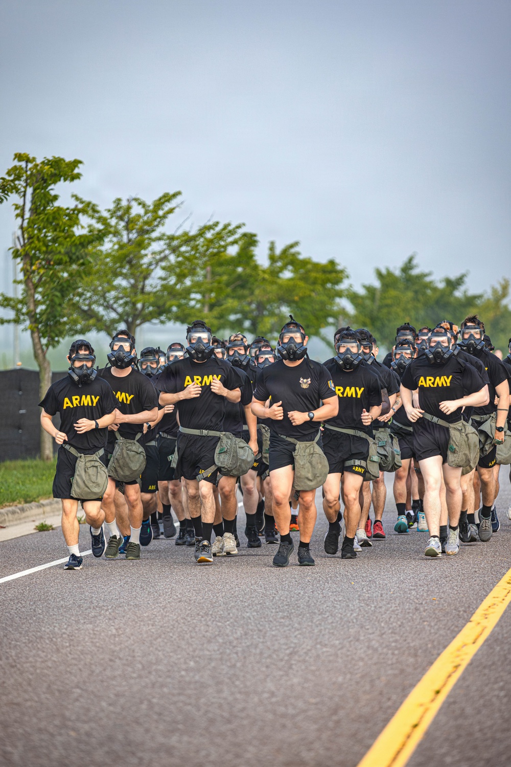 A large group of U.S. Army soldiers are running in formation along a paved road while wearing gas masks and carrying what appear to be gas mask carrier pouches on their belts. They are dressed in standard Army PT (Physical Training) uniforms consisting of black 'ARMY' t-shirts, olive/khaki shorts, and various athletic footwear. This appears to be a military physical training exercise or gas mask run drill conducted in an outdoor setting.