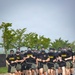 A large group of U.S. Army soldiers are running in formation along a paved road while wearing gas masks and carrying what appear to be gas mask carrier pouches on their belts. They are dressed in standard Army PT (Physical Training) uniforms consisting of black 'ARMY' t-shirts, olive/khaki shorts, and various athletic footwear. This appears to be a military physical training exercise or gas mask run drill conducted in an outdoor setting.