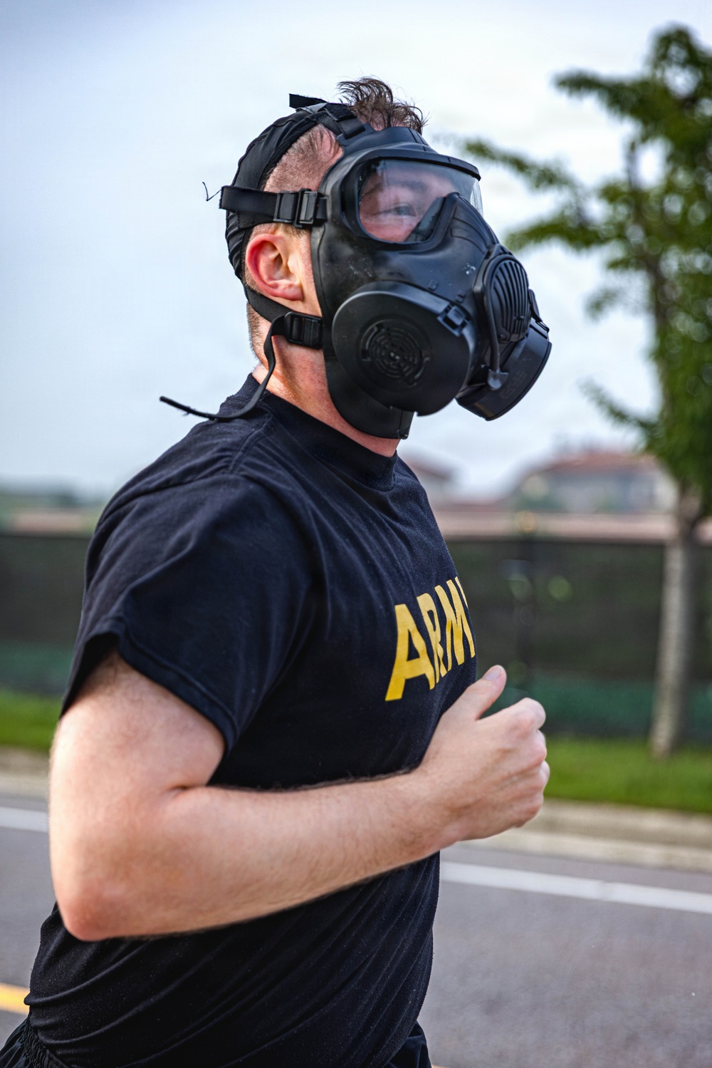 A male individual wearing a black U.S. Army physical training t-shirt and a black military-style gas mask is running on a paved road or track. The image appears to depict a military fitness training exercise involving CBRN (Chemical, Biological, Radiological, Nuclear) protective equipment. The background shows a blurred outdoor environment with trees and structures.