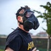 A male individual wearing a black U.S. Army physical training t-shirt and a black military-style gas mask is running on a paved road or track. The image appears to depict a military fitness training exercise involving CBRN (Chemical, Biological, Radiological, Nuclear) protective equipment. The background shows a blurred outdoor environment with trees and structures.