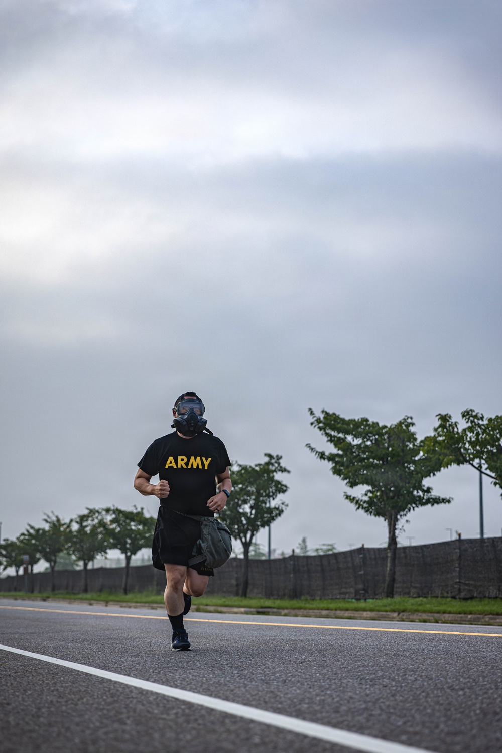 A soldier wearing a black U.S. Army PT (physical training) shirt and a gas mask is running along a paved road while carrying a dark duffel bag. The setting appears to be a military installation or base, with trees, a perimeter wall, and an overcast sky in the background. The image captures what appears to be a military fitness or training exercise.