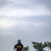 A soldier wearing a black U.S. Army PT (physical training) shirt and a gas mask is running along a paved road while carrying a dark duffel bag. The setting appears to be a military installation or base, with trees, a perimeter wall, and an overcast sky in the background. The image captures what appears to be a military fitness or training exercise.