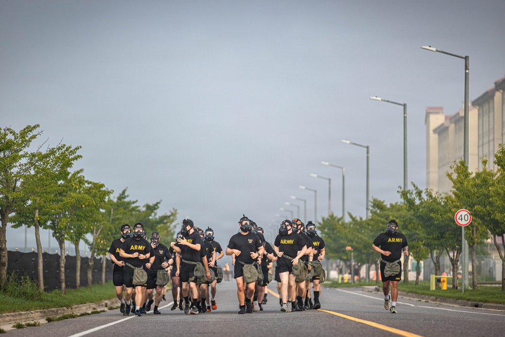 A group of U.S. Army personnel are running in formation along a wide road while wearing gas masks and black PT (physical training) uniforms. Many of the runners are carrying olive-drab military bags. The setting appears to be a military installation, indicated by the structured road, street lighting, and surrounding buildings.