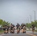A group of U.S. Army personnel are running in formation along a wide road while wearing gas masks and black PT (physical training) uniforms. Many of the runners are carrying olive-drab military bags. The setting appears to be a military installation, indicated by the structured road, street lighting, and surrounding buildings.