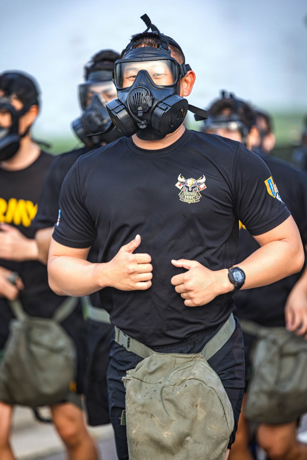 A group of military personnel are running while wearing gas masks and black t-shirts. The lead figure wears a black t-shirt with a 'Vikings HHC' unit patch and a military shoulder sleeve insignia, along with olive drab shorts and a tactical belt. The setting appears to be an outdoor physical training exercise or formation run.