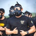 A group of military personnel are running while wearing gas masks and black t-shirts. The lead figure wears a black t-shirt with a 'Vikings HHC' unit patch and a military shoulder sleeve insignia, along with olive drab shorts and a tactical belt. The setting appears to be an outdoor physical training exercise or formation run.