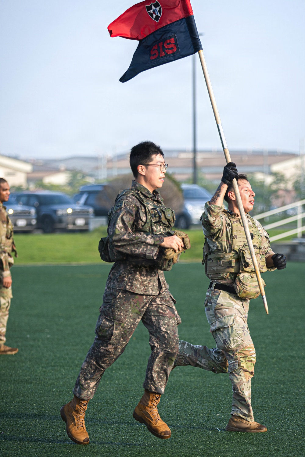 Two soldiers in different military uniforms are running across a green turf field, with one carrying a unit guidon flag. The flag is red and navy blue, bearing the text 'SIS' and what appears to be a 2nd Infantry Division patch. The setting appears to be a military installation, likely in South Korea based on the Korean Army (ROKA) uniform worn by one soldier.