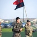 Two soldiers in different military uniforms are running across a green turf field, with one carrying a unit guidon flag. The flag is red and navy blue, bearing the text 'SIS' and what appears to be a 2nd Infantry Division patch. The setting appears to be a military installation, likely in South Korea based on the Korean Army (ROKA) uniform worn by one soldier.