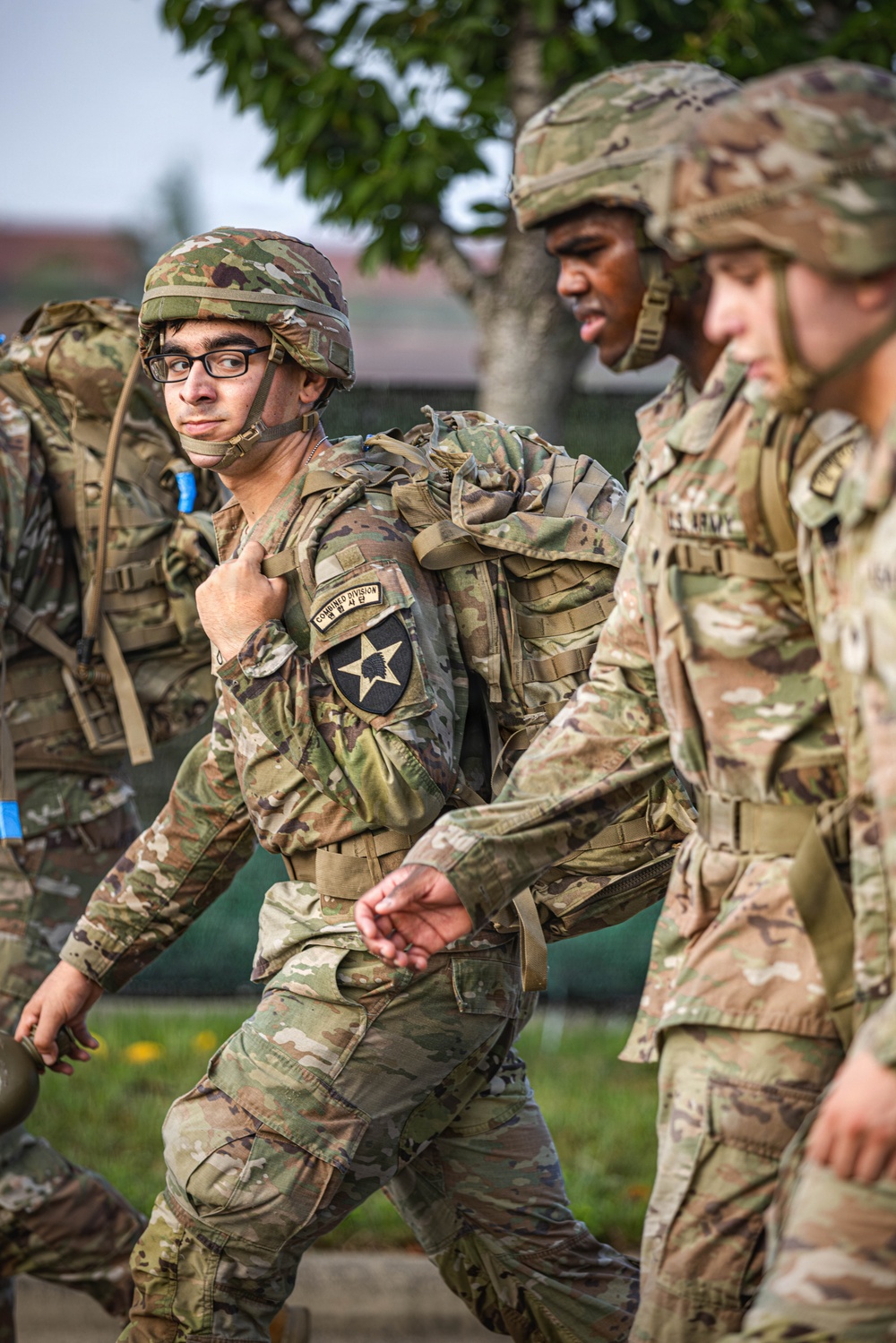 A group of U.S. Army soldiers in Operational Camouflage Pattern (OCP) uniforms are conducting a ruck march or physical training exercise while carrying large rucksacks. The central soldier, wearing glasses and a combat helmet, displays a '2nd Infantry Division / Combined Division' shoulder patch, indicating assignment to a unit stationed in South Korea. The soldiers appear to be moving at a brisk pace in an outdoor military installation setting.