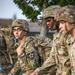 A group of U.S. Army soldiers in Operational Camouflage Pattern (OCP) uniforms are conducting a ruck march or physical training exercise while carrying large rucksacks. The central soldier, wearing glasses and a combat helmet, displays a '2nd Infantry Division / Combined Division' shoulder patch, indicating assignment to a unit stationed in South Korea. The soldiers appear to be moving at a brisk pace in an outdoor military installation setting.