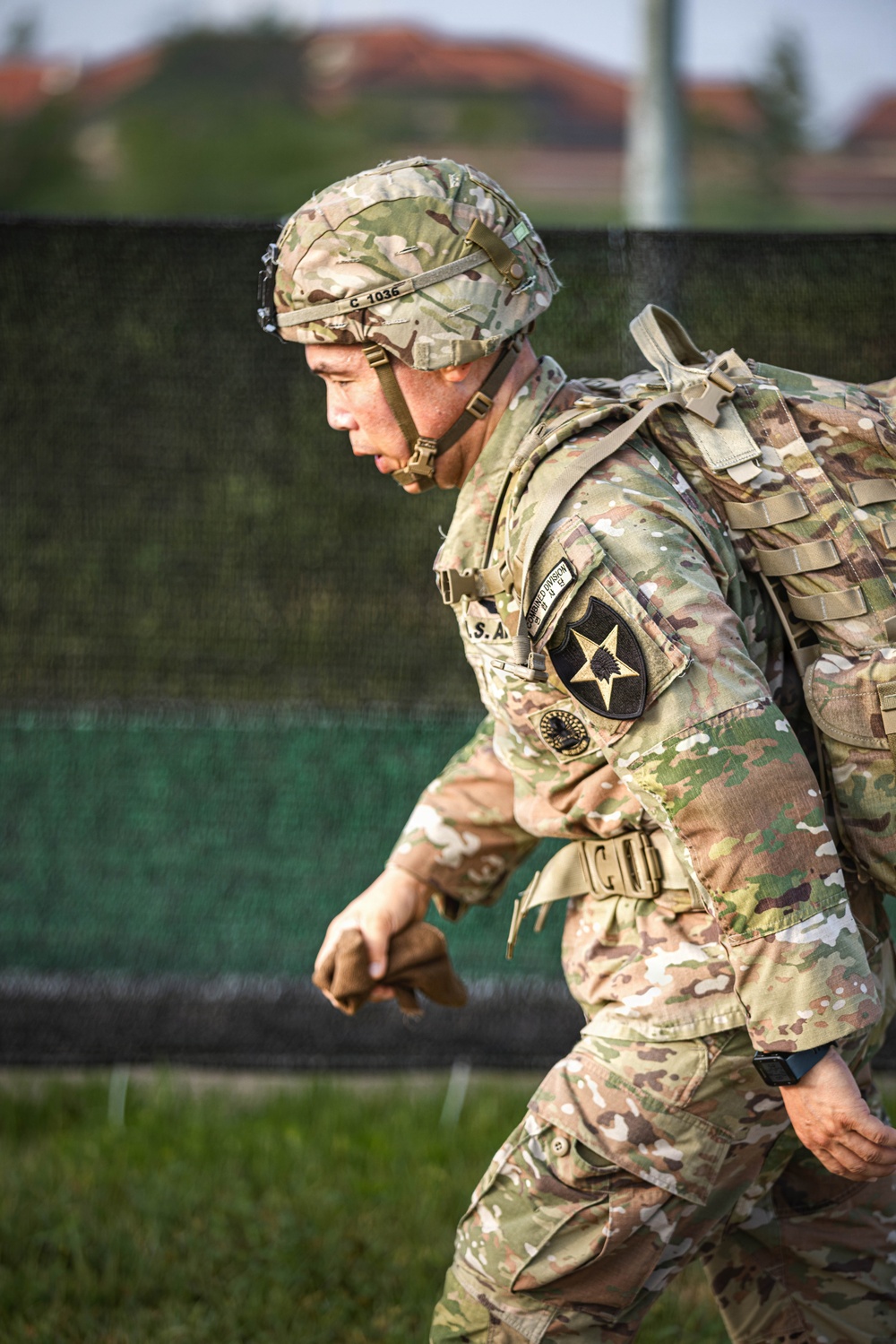 A U.S. Army soldier in full OCP (Operational Camouflage Pattern) uniform is moving forward under apparent rain conditions, leaning forward under the weight of a rucksack. The soldier is wearing a helmet marked 'C 1035' and displays a 2nd Infantry Division shoulder sleeve insignia patch. The image appears to capture a physical fitness event or ruck march exercise.