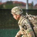 A U.S. Army soldier in full OCP (Operational Camouflage Pattern) uniform is moving forward under apparent rain conditions, leaning forward under the weight of a rucksack. The soldier is wearing a helmet marked 'C 1035' and displays a 2nd Infantry Division shoulder sleeve insignia patch. The image appears to capture a physical fitness event or ruck march exercise.