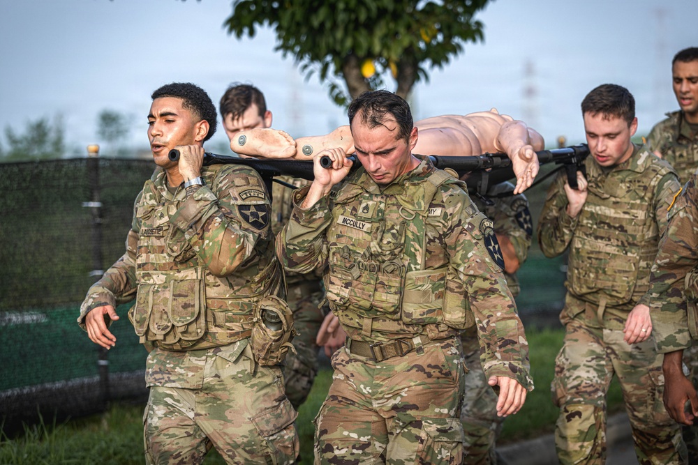 A group of U.S. Army soldiers in MultiCam/OCP camouflage uniforms are conducting a litter/casualty carry drill, transporting a training mannequin on a stretcher pole. The two lead soldiers, identified by name tapes as 'LAUSSETTE' and 'McCULLY', are bearing the weight of the simulated casualty on their shoulders. This appears to be a combat readiness or medical evacuation training exercise.