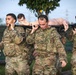 A group of U.S. Army soldiers in MultiCam/OCP camouflage uniforms are conducting a litter/casualty carry drill, transporting a training mannequin on a stretcher pole. The two lead soldiers, identified by name tapes as 'LAUSSETTE' and 'McCULLY', are bearing the weight of the simulated casualty on their shoulders. This appears to be a combat readiness or medical evacuation training exercise.