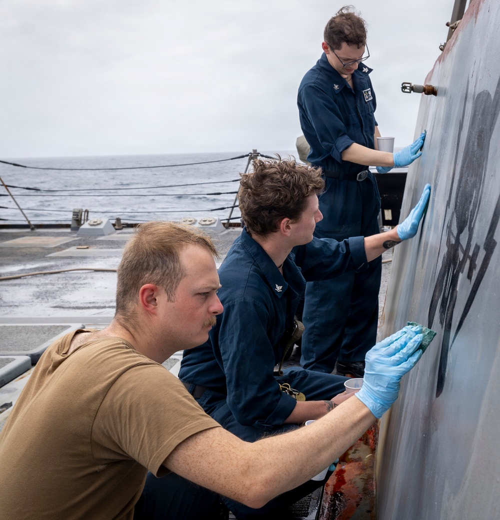 Three naval personnel are performing maintenance work on the exterior hull or superstructure of a naval vessel at sea. Two individuals are wearing navy blue coveralls (utility uniforms) while one wears a tan/coyote brown t-shirt, all wearing blue nitrile gloves and using cloths or abrasive pads to clean or treat a metal surface. The overcast sky and visible ocean in the background confirm the ship is underway.