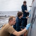 Three naval personnel are performing maintenance work on the exterior hull or superstructure of a naval vessel at sea. Two individuals are wearing navy blue coveralls (utility uniforms) while one wears a tan/coyote brown t-shirt, all wearing blue nitrile gloves and using cloths or abrasive pads to clean or treat a metal surface. The overcast sky and visible ocean in the background confirm the ship is underway.