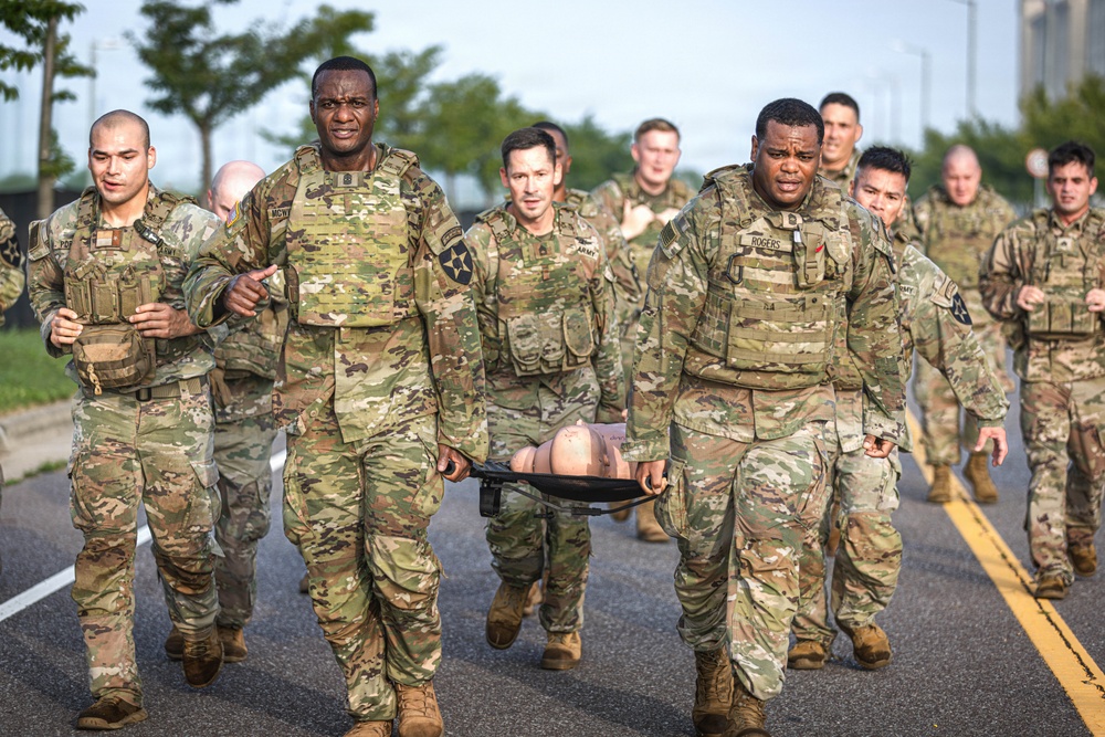 A group of U.S. Army soldiers in OCP (Operational Camouflage Pattern) uniforms are conducting what appears to be a medical training exercise on a road. Two soldiers in the foreground are carrying a stretcher with what appears to be a training mannequin or simulated casualty. A large group of soldiers follows behind, suggesting a unit-level training event or physical readiness exercise.