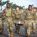 A group of U.S. Army soldiers in OCP (Operational Camouflage Pattern) uniforms are conducting what appears to be a medical training exercise on a road. Two soldiers in the foreground are carrying a stretcher with what appears to be a training mannequin or simulated casualty. A large group of soldiers follows behind, suggesting a unit-level training event or physical readiness exercise.