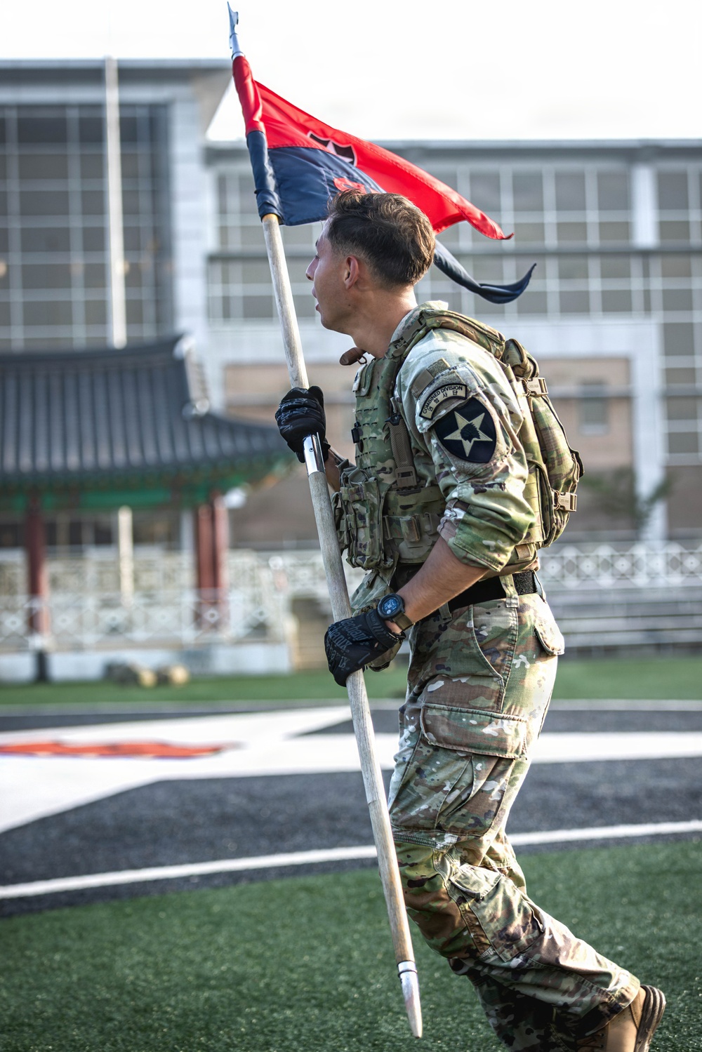 A U.S. Army soldier in full combat gear carries a military guidon flag while moving across what appears to be an athletic field or parade ground. The soldier wears a 2nd Infantry Division patch on his left shoulder, suggesting this is likely taking place in South Korea. The background includes what appears to be a traditional Korean-style structure alongside modern buildings.