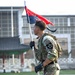 A U.S. Army soldier in full combat gear carries a military guidon flag while moving across what appears to be an athletic field or parade ground. The soldier wears a 2nd Infantry Division patch on his left shoulder, suggesting this is likely taking place in South Korea. The background includes what appears to be a traditional Korean-style structure alongside modern buildings.