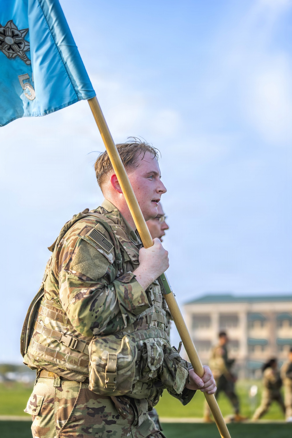A U.S. Army soldier in OCP (Operational Camouflage Pattern) uniform carries a light blue guidon flag with the number '5' and a military branch insignia during what appears to be a formation run or physical training event. The soldier is wearing a rucksack and tactical gear, suggesting this may be a ruck march or similar endurance event. Additional soldiers in uniform are visible in the blurred background near a military building.