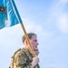 A U.S. Army soldier in OCP (Operational Camouflage Pattern) uniform carries a light blue guidon flag with the number '5' and a military branch insignia during what appears to be a formation run or physical training event. The soldier is wearing a rucksack and tactical gear, suggesting this may be a ruck march or similar endurance event. Additional soldiers in uniform are visible in the blurred background near a military building.