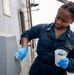 A U.S. Navy sailor wearing dark blue utility coveralls is performing maintenance work on the exterior of a naval vessel. She is applying paint or a surface treatment to a gray bulkhead wall while holding a white cup containing paint and a brush. The work is being conducted on the open deck of the ship with ocean visible in the background.