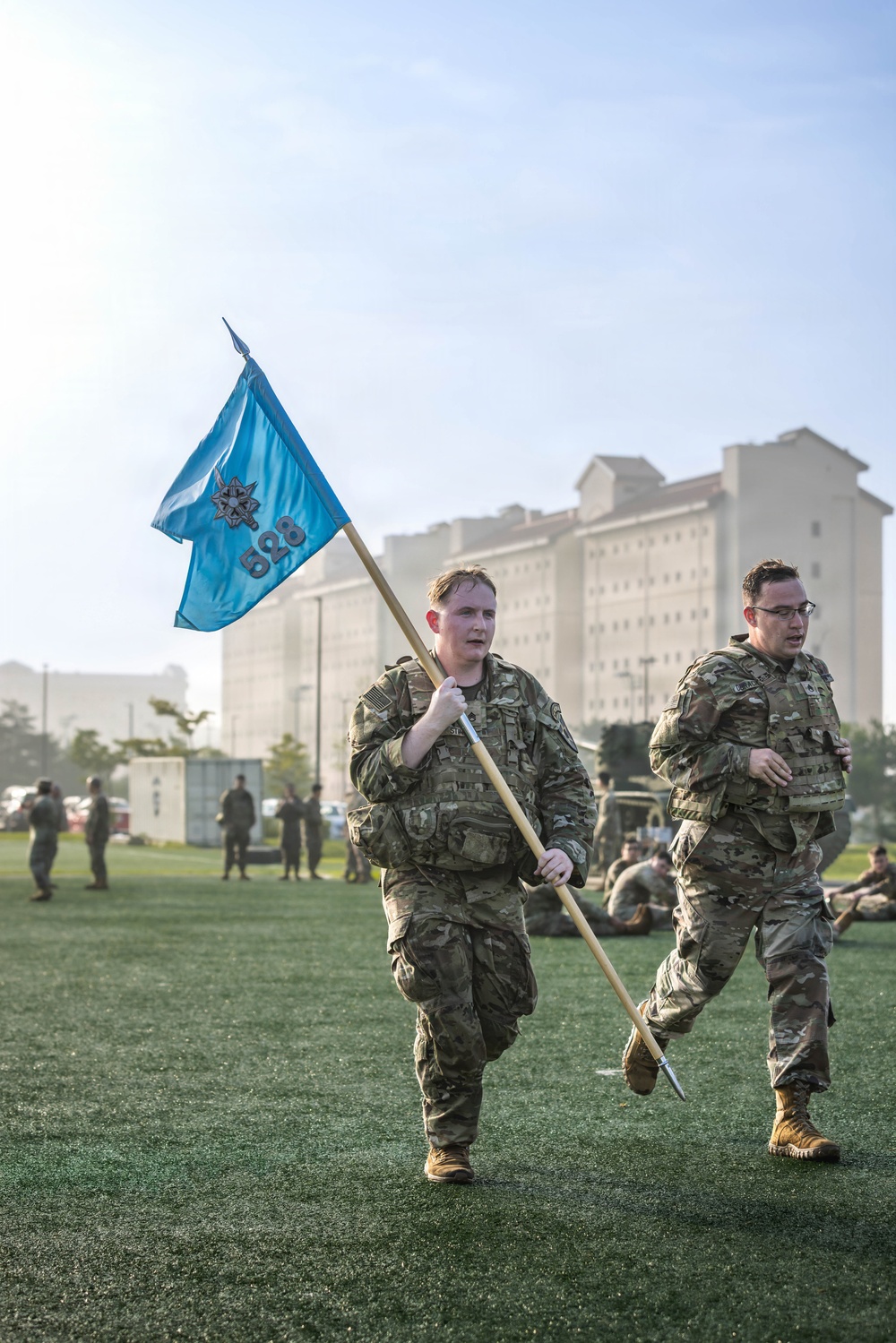 Two U.S. Army soldiers in OCP (Operational Camouflage Pattern) uniforms are running across a green turf field, with the lead soldier carrying a light blue guidon flag bearing the number '528' and a military insignia. Additional soldiers are visible in the background, some resting on the ground and others standing near military vehicles and shipping containers. The setting appears to be a military installation, likely during a physical training event or unit formation activity.