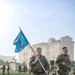 Two U.S. Army soldiers in OCP (Operational Camouflage Pattern) uniforms are running across a green turf field, with the lead soldier carrying a light blue guidon flag bearing the number '528' and a military insignia. Additional soldiers are visible in the background, some resting on the ground and others standing near military vehicles and shipping containers. The setting appears to be a military installation, likely during a physical training event or unit formation activity.