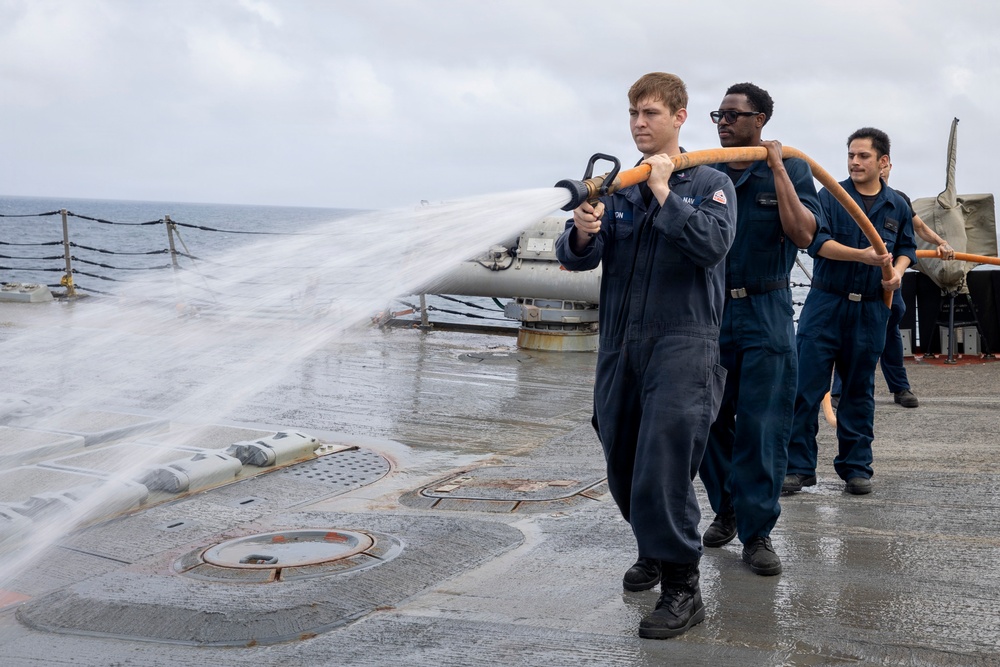 Three U.S. Navy sailors in dark blue coveralls are conducting a fire hose drill on the wet deck of a naval vessel at sea. The lead sailor is operating a large fire hose nozzle, directing a powerful stream of water across the ship's deck. The overcast sky and open ocean are visible in the background, suggesting the exercise is taking place underway.