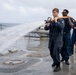 Three U.S. Navy sailors in dark blue coveralls are conducting a fire hose drill on the wet deck of a naval vessel at sea. The lead sailor is operating a large fire hose nozzle, directing a powerful stream of water across the ship's deck. The overcast sky and open ocean are visible in the background, suggesting the exercise is taking place underway.