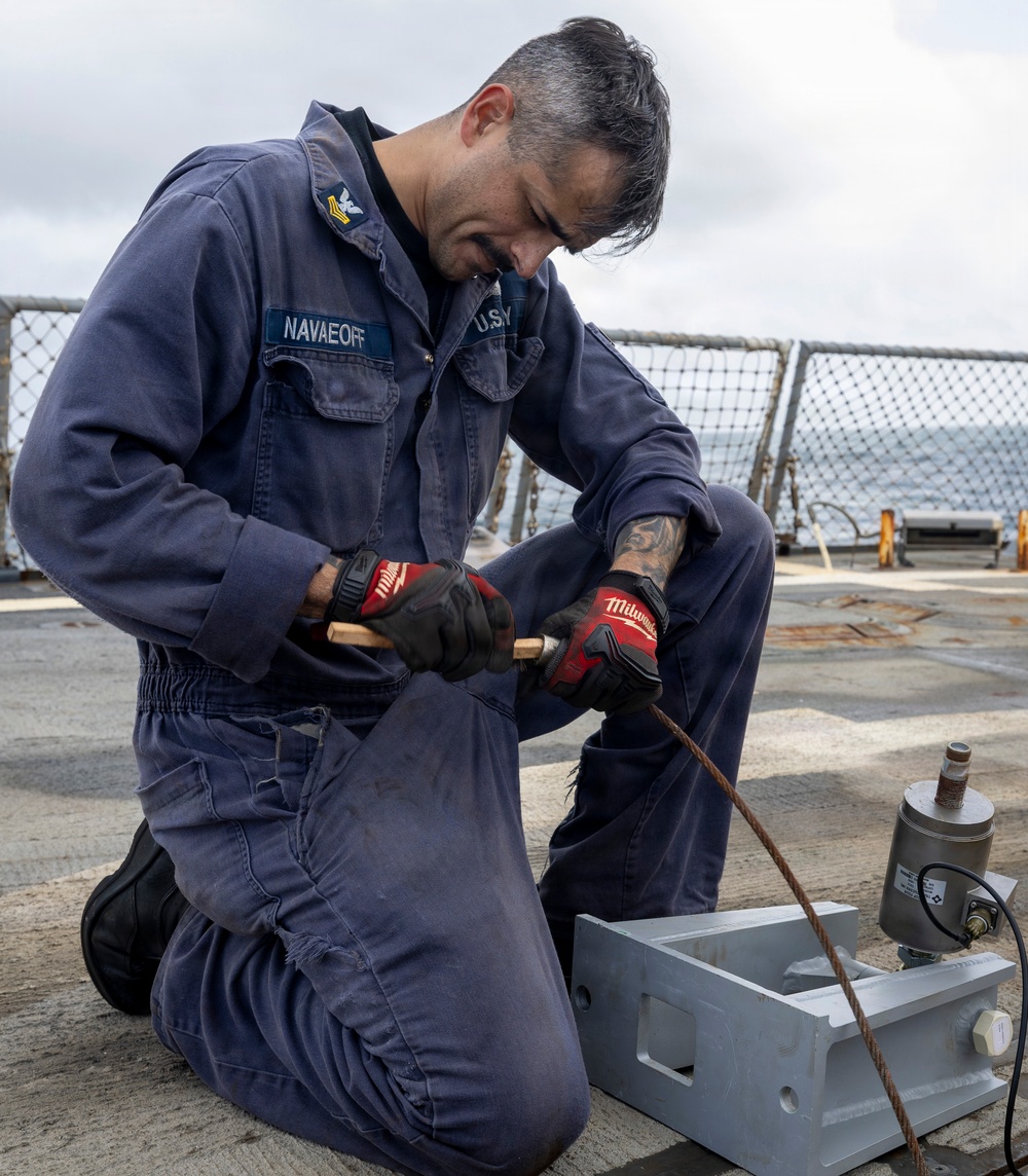 A U.S. Navy sailor identified by name tape as 'NAVAEOFF' is kneeling on the deck of a naval vessel performing maintenance work. The sailor is wearing blue coveralls (utilities/coveralls), Milwaukee brand work gloves, and is working with a metal component and wire/cable. The setting is an open ship deck with safety netting and ocean visible in the background.