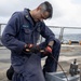 A U.S. Navy sailor identified by name tape as 'NAVAEOFF' is kneeling on the deck of a naval vessel performing maintenance work. The sailor is wearing blue coveralls (utilities/coveralls), Milwaukee brand work gloves, and is working with a metal component and wire/cable. The setting is an open ship deck with safety netting and ocean visible in the background.