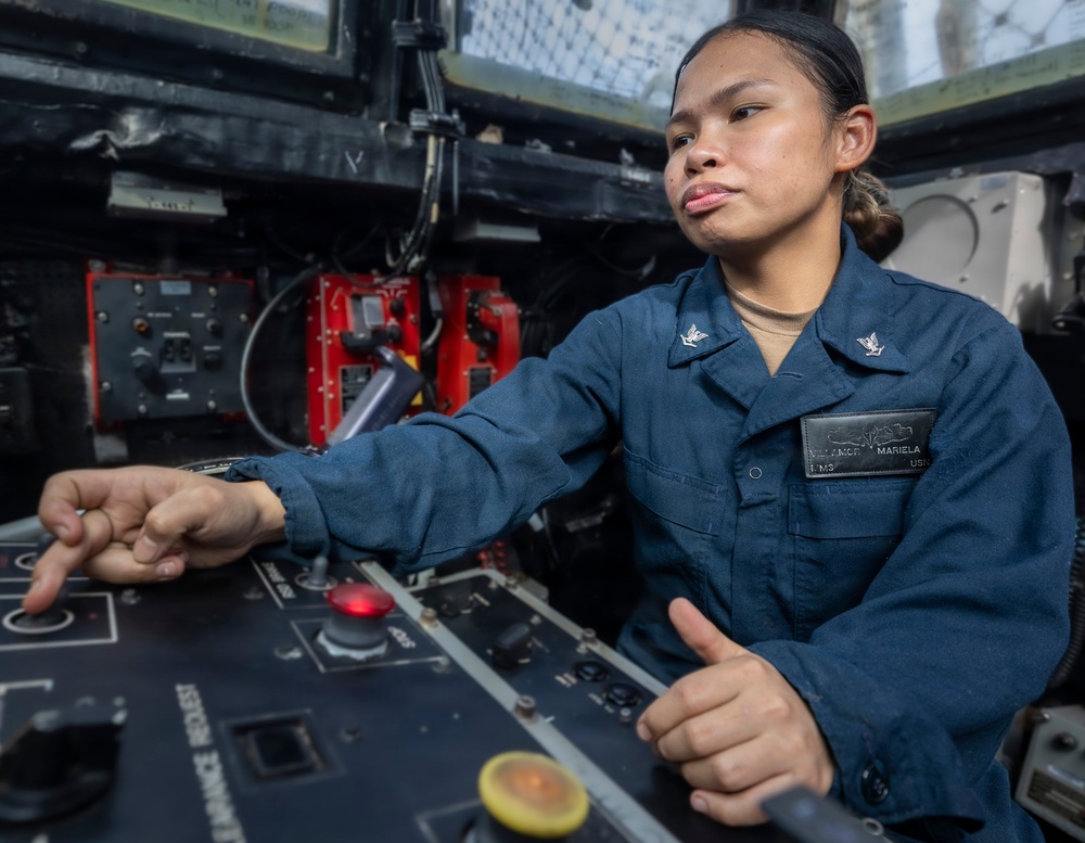 A U.S. Navy sailor, identified by name tag as 'Mariela' with last name partially visible as 'Villamore' or similar, is operating a control panel aboard a naval vessel. She is wearing a dark blue Navy working uniform with petty officer collar insignia and a USN name tag. The image captures her in an active operational environment with various switches, indicators, and equipment visible.
