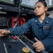 A U.S. Navy sailor, identified by name tag as 'Mariela' with last name partially visible as 'Villamore' or similar, is operating a control panel aboard a naval vessel. She is wearing a dark blue Navy working uniform with petty officer collar insignia and a USN name tag. The image captures her in an active operational environment with various switches, indicators, and equipment visible.
